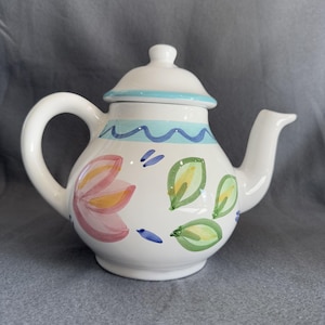 May include: A white ceramic teapot with a floral design. The teapot has a light blue lid with a wavy blue and white trim. The floral design features pink, yellow, and green flowers and leaves.
