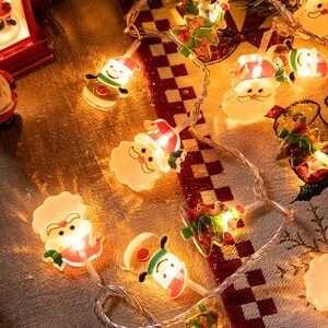 May include: A string of Christmas lights featuring a variety of characters, including Santa Claus, a snowman, and a Christmas tree. The lights are white and the characters are colourful.