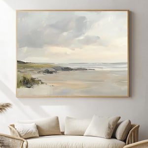 May include: A framed landscape painting depicts a serene beach scene with a cloudy sky. The artwork features soft, muted tones of beige, gray, and green, creating a calming atmosphere. The painting is displayed above a beige sofa and wicker furniture.