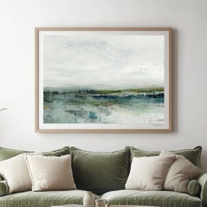 May include: A framed abstract landscape painting hangs above a green velvet sofa with cream cushions. The artwork depicts a tranquil scene in muted blues, greens, and whites. A round wooden coffee table is positioned in front of the sofa, creating a cosy living space.