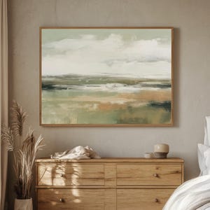 Puede incluir: Pintura abstracta de paisaje en un marco de madera. La obra presenta tonos apagados de verde, beige y marrón, que representan una escena serena con un cielo nublado y una masa de agua. La pintura cuelga sobre una cómoda de madera.