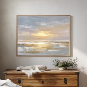 Puede incluir: Pintura abstracta enmarcada con marco de madera. La obra representa una puesta de sol sobre el agua, con tonos dorados, azules y blancos. La pintura cuelga sobre un cofre de madera, con una manta de color neutro y objetos decorativos.