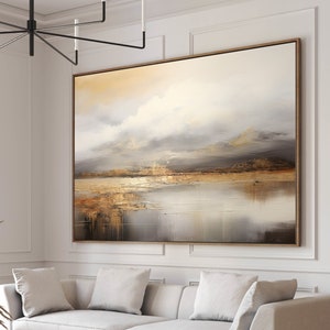 May include: A white sofa with three cushions sits in front of a large abstract landscape painting with muted tones of brown, beige, and grey. The painting is framed in a thin, gold frame. A black coffee table with a wooden tray and books sits in front of the sofa.