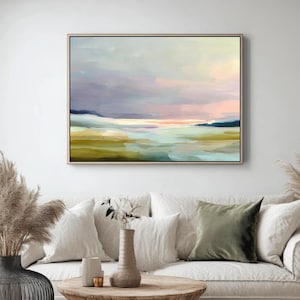 May include: Abstract landscape painting in a wooden frame. The artwork features a serene scene with a blend of colors, including shades of green, blue, pink, and purple. The painting is displayed above a white sofa with decorative pillows and a wooden coffee table.