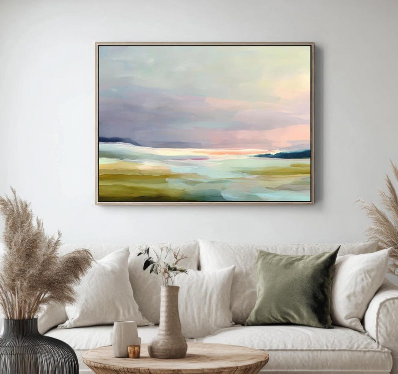 May include: Abstract landscape painting in a wooden frame. The artwork features a serene scene with a blend of colors, including shades of green, blue, pink, and purple. The painting is displayed above a white sofa with decorative pillows and a wooden coffee table.