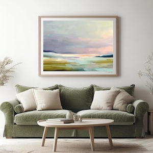 May include: Abstract landscape art print in a wooden frame. The artwork features a serene scene with pastel colors, including blues, pinks, and greens, evoking a sense of calm. The painting is displayed above a green sofa with beige pillows.