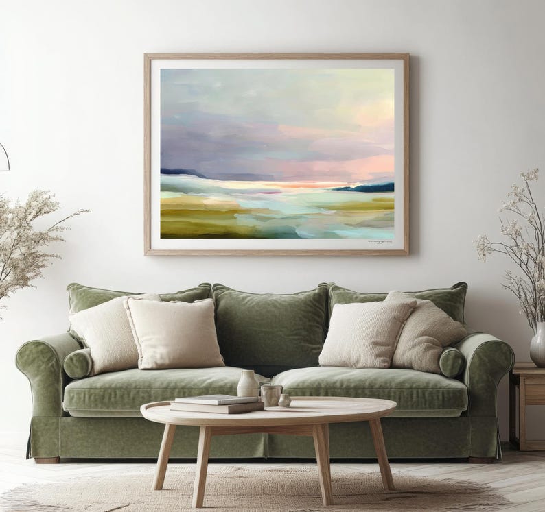 May include: Abstract landscape art print in a wooden frame. The artwork features a serene scene with pastel colors, including blues, pinks, and greens, evoking a sense of calm. The painting is displayed above a green sofa with beige pillows.