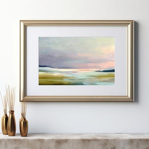 May include: An abstract landscape painting in a gold frame. The artwork features a soft color palette of blues, greens, and pinks, depicting a serene scene of a body of water and distant hills. The painting is displayed on a white wall.