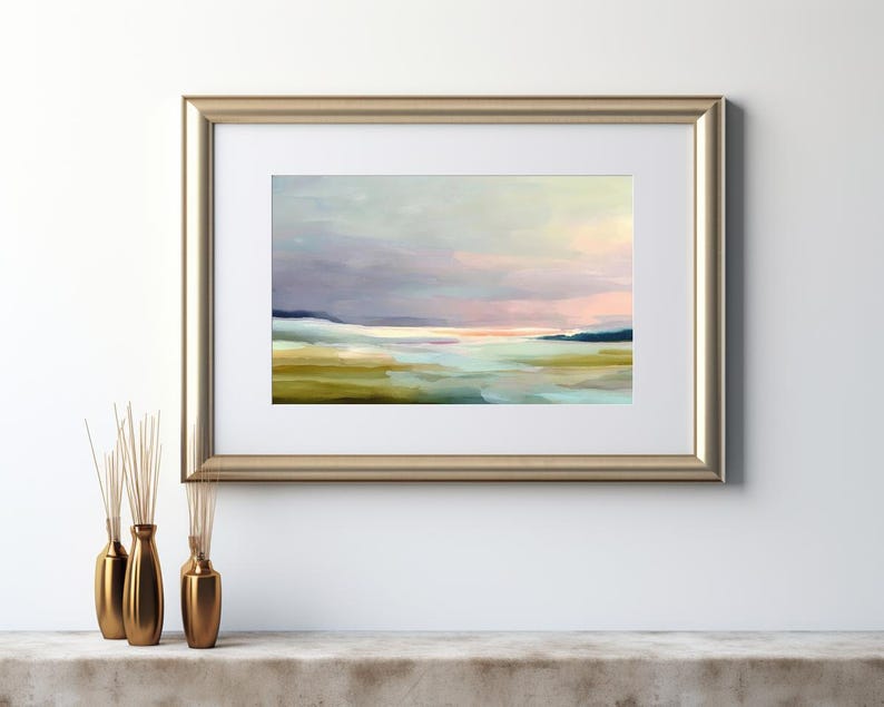 May include: An abstract landscape painting in a gold frame. The artwork features a soft color palette of blues, greens, and pinks, depicting a serene scene of a body of water and distant hills. The painting is displayed on a white wall.