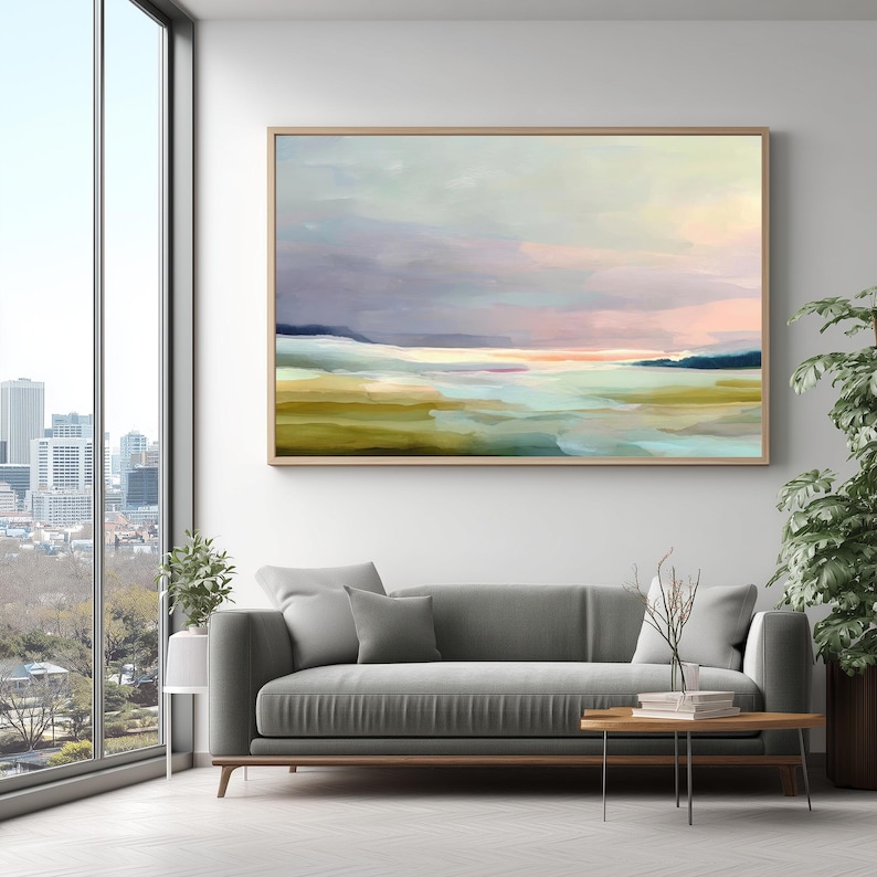 May include: Abstract landscape painting in a wooden frame. The artwork features a serene scene with a blend of colors, including blues, greens, and pinks, evoking a sense of calm. The painting is displayed above a gray sofa.