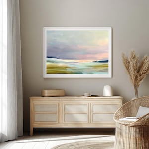 May include: A framed abstract landscape painting with a white border. The artwork features a serene scene with pastel colors, including blues, greens, and pinks, evoking a sense of calm. The painting is displayed above a wooden cabinet with woven panels.