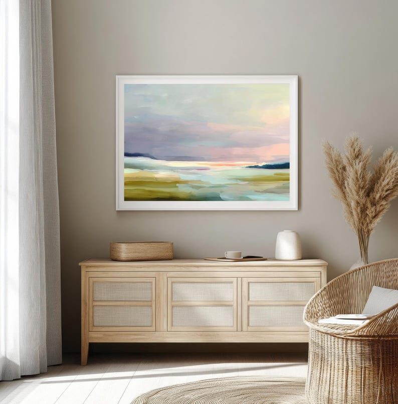 May include: A framed abstract landscape painting with a white border. The artwork features a serene scene with pastel colors, including blues, greens, and pinks, evoking a sense of calm. The painting is displayed above a wooden cabinet with woven panels.