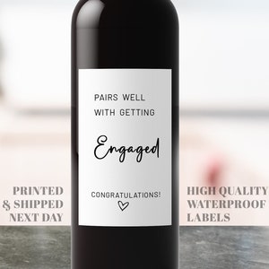 May include: A wine bottle with a white label that says "Pairs well with getting engaged" and "Congratulations!" with a heart. The label also says "Printed & Shipped Next Day" and "High Quality Waterproof Labels".