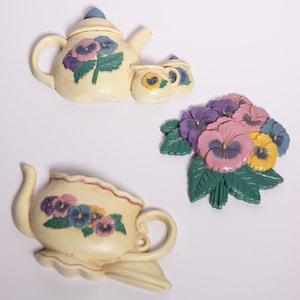 Pode incluir: Decoração de parede creme com uma chaleira, uma chávena e um buquê de amores-perfeitos. A chaleira e a chávena são adornadas com desenhos florais de amores-perfeitos em roxo, rosa e azul. O buquê tem folhas verdes.