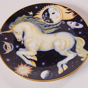 Puede incluir: Plato decorativo con un unicornio blanco, cuerno dorado y fondo negro lleno de estrellas, planetas y cuerpos celestes. El plato tiene un borde dorado y un diseño de sol, luna y estrellas.
