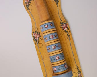 Puede incluir: Rodillo de madera con mango marrón claro y base decorativa. La base está pintada de azul claro con diseños florales. La tabla de madera es de color amarillo claro con rosas rosas y hojas verdes. La tabla mide 30 cm de largo.