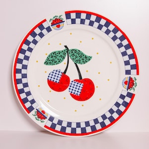 May include: White ceramic plate with a red and blue chequered border. The plate features a central design of two red cherries with blue chequered patches, green leaves, and small yellow dots. Additional cherry illustrations are on the border.