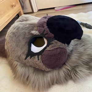May include: A plush animal head mask featuring long, shaggy grey faux fur, a prominent black snout, and dark purple accents around the eyes and nose area. The mask includes large, stylized black eye frames with white inner eye openings.