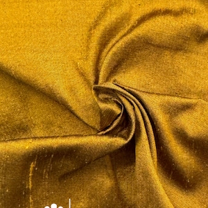 May include: Close-up of a golden-yellow fabric with a subtle sheen, showcasing intricate folds and textures. The fabric's surface reflects light, creating highlights and shadows. The logo "marce and emma" is visible in the lower right corner.