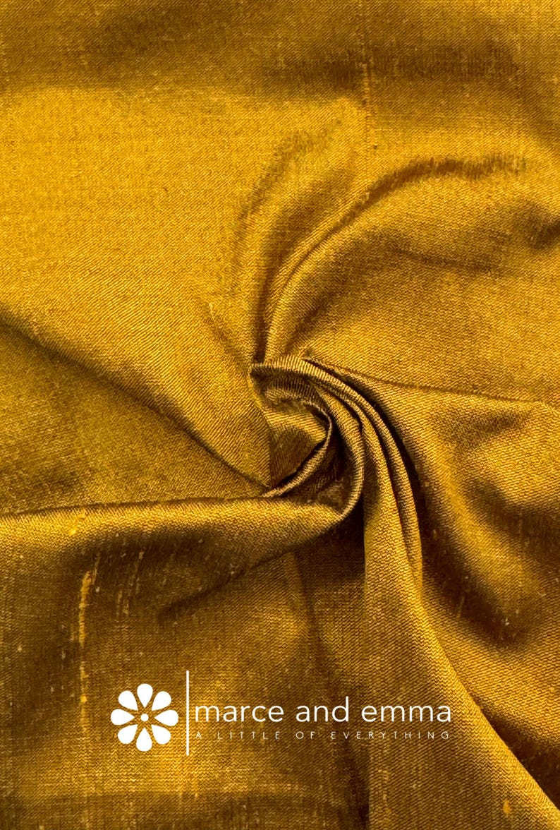 May include: Close-up of a golden-yellow fabric with a subtle sheen, showcasing intricate folds and textures. The fabric's surface reflects light, creating highlights and shadows. The logo "marce and emma" is visible in the lower right corner.