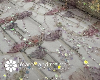 May include: Close-up of a fabric with a floral design. The fabric features embroidered flowers in shades of purple and pink, accented with silver beads and iridescent sequins. The text "marce and emma a little of everything" is visible.