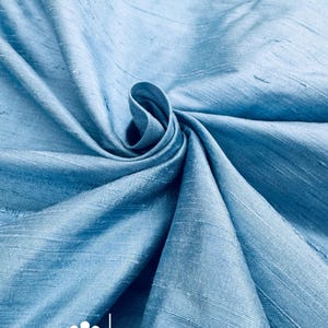 May include: Light blue fabric with a subtle sheen, artfully draped and swirled. The fabric's texture is visible, with a soft, flowing appearance. The text "marce and emma" is displayed with a floral logo.