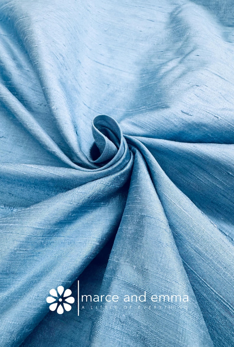 May include: Light blue fabric with a subtle sheen, artfully draped and swirled. The fabric's texture is visible, with a soft, flowing appearance. The text "marce and emma" is displayed with a floral logo.