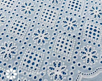 May include: Close-up of a light blue fabric with intricate white embroidery. The design features floral and geometric patterns, including flowers and dotted squares. The fabric has a scalloped edge and the text "marce and emma" is visible.