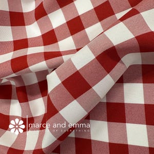 May include: Close-up of red and white checkered fabric. The fabric is folded, showing the texture and pattern. The fabric has a classic gingham design, with large squares of red and white. The brand name "marce and emma" is visible.