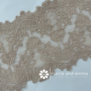 May include: Close-up of a beige lace trim with an intricate floral and scroll design. The lace features a delicate, openwork pattern with detailed edges. The trim is likely used for sewing, crafting, or adding embellishments to garments or home decor.