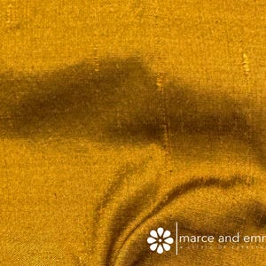 May include: Close-up of a golden-yellow fabric with visible texture and subtle variations in tone. The material appears to be draped, with soft folds and shadows adding depth. The fabric has a slight sheen, suggesting a luxurious texture.