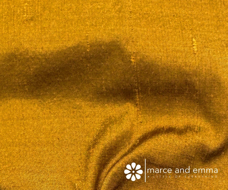 May include: Close-up of a golden-yellow fabric with visible texture and subtle variations in tone. The material appears to be draped, with soft folds and shadows adding depth. The fabric has a slight sheen, suggesting a luxurious texture.
