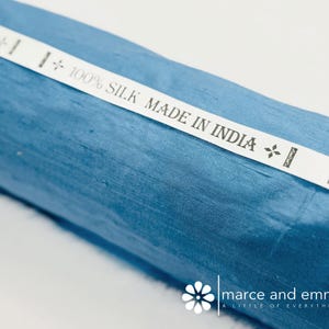 May include: A roll of blue silk fabric with a white label that reads "100% SILK MADE IN INDIA." The fabric has a subtle sheen and a textured appearance. The label has decorative floral accents.