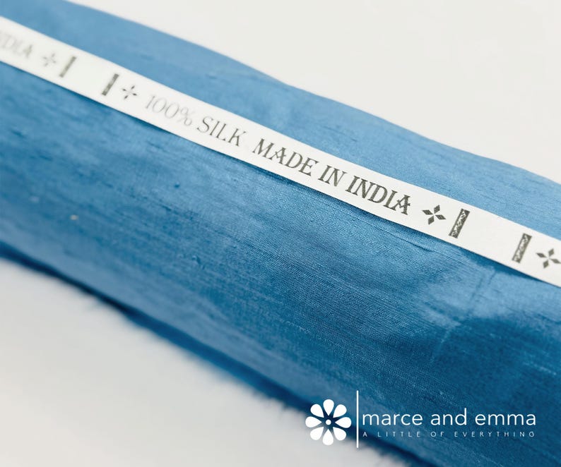 May include: A roll of blue silk fabric with a white label that reads "100% SILK MADE IN INDIA." The fabric has a subtle sheen and a textured appearance. The label has decorative floral accents.