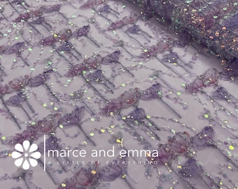 May include: A roll of sheer fabric with a floral pattern in shades of purple and lavender. The fabric is embellished with iridescent sequins, adding a shimmering effect. The brand name "marce and emma" is visible.