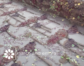 May include: Close-up of a delicate fabric with floral embroidery and iridescent sequins. The design features purple and pink flowers with beaded stems. The fabric is semi-sheer, with the text "marce and emma a little of everything" in the lower left corner.