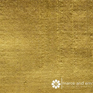 May include: Close-up of a textured, golden-yellow fabric. The weave is visible, with vertical lines creating a subtle striped effect. The fabric has a slightly rough appearance. The logo "marce and emma" is in the bottom right corner.