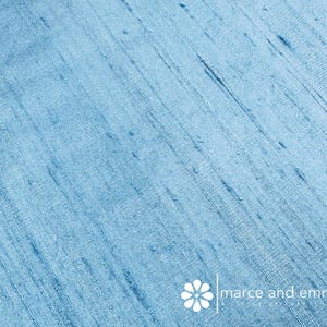 May include: Close-up of a light blue fabric with a textured, diagonal weave. The fabric has subtle variations in color and texture, with darker blue streaks. The text "marce and emma" is visible in the lower right corner.