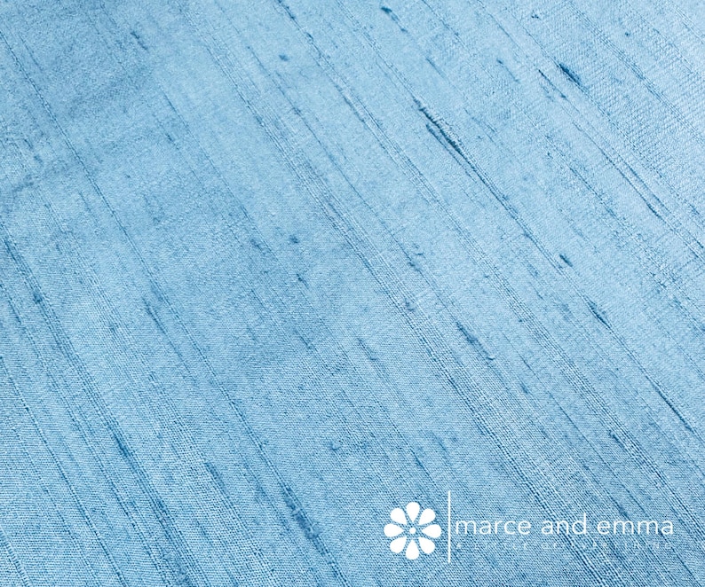 May include: Close-up of a light blue fabric with a textured, diagonal weave. The fabric has subtle variations in color and texture, with darker blue streaks. The text "marce and emma" is visible in the lower right corner.
