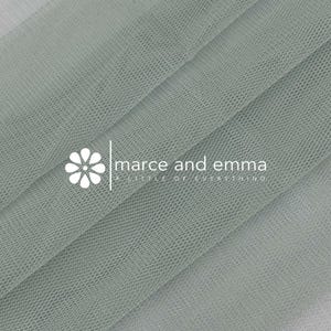May include: Close-up of a folded, light green tulle fabric. The fabric has a fine mesh texture and is layered, creating shadows and depth. The text "marce and emma a little of everything" is printed on the fabric.