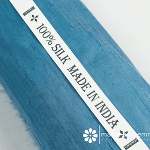 May include: Close-up of a roll of teal-colored silk fabric. A white label with the text "100% SILK MADE IN INDIA" is diagonally across the fabric. The fabric has a textured appearance. The label also includes a floral design and the brand name.
