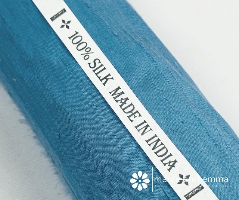 May include: Close-up of a roll of teal-colored silk fabric. A white label with the text "100% SILK MADE IN INDIA" is diagonally across the fabric. The fabric has a textured appearance. The label also includes a floral design and the brand name.