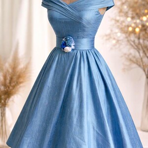 May include: A light blue, off-the-shoulder dress with a fitted bodice and a full skirt. The dress features a decorative floral accent at the waist. The fabric has a subtle sheen, and the dress is displayed on a mannequin.