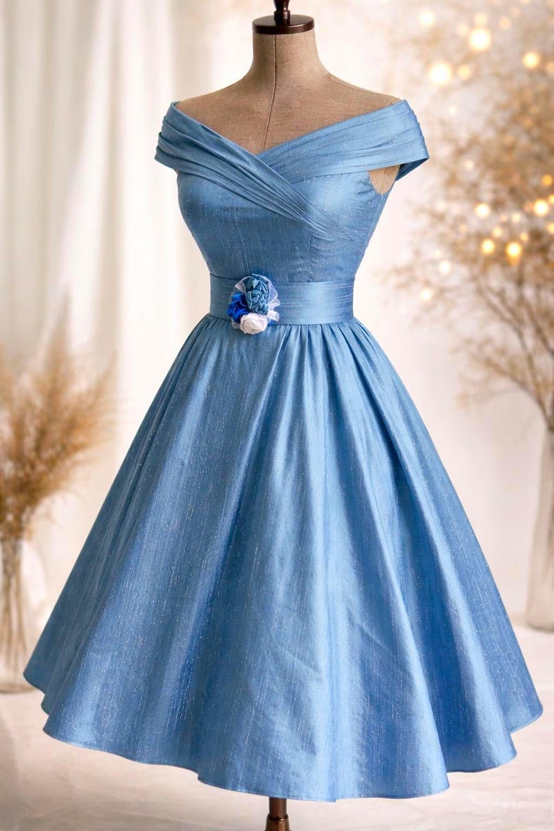 May include: A light blue, off-the-shoulder dress with a fitted bodice and a full skirt. The dress features a decorative floral accent at the waist. The fabric has a subtle sheen, and the dress is displayed on a mannequin.