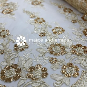 May include: Ivory lace fabric with intricate gold floral embroidery. The design features detailed flowers, leaves, and vines, with a subtle sheen. The fabric is likely used for bridal wear or special occasion garments. The text "marce and emma" is visible.