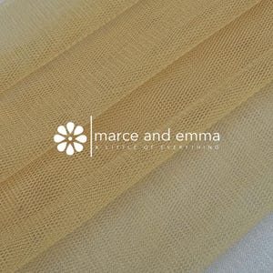 May include: Close-up of a folded, sheer fabric in shades of gold and tan. The fabric has a fine, net-like texture. The text "marce and emma a little of everything" is printed on the fabric.