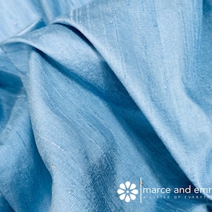 May include: Close-up of a light blue fabric with visible texture and folds. The fabric has a subtle sheen, suggesting a silk or similar material. The image is well-lit, highlighting the color and texture of the material. The fabric is likely used for clothing or home decor.