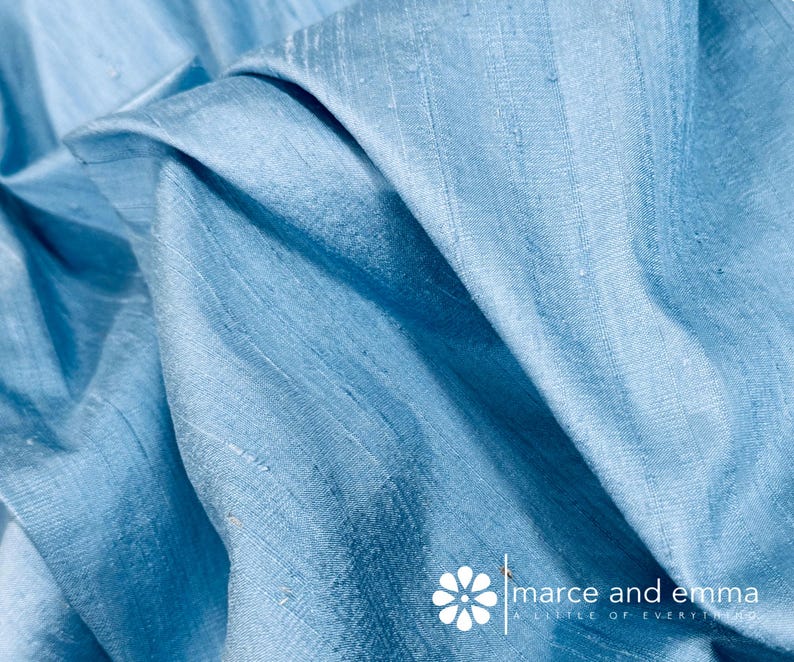 May include: Close-up of a light blue fabric with visible texture and folds. The fabric has a subtle sheen, suggesting a silk or similar material. The image is well-lit, highlighting the color and texture of the material. The fabric is likely used for clothing or home decor.