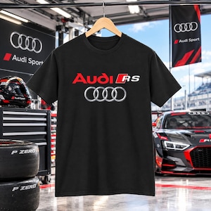 May include: A black short-sleeve t-shirt featuring the red and white Audi RS logo with the iconic four silver rings emblem centered on the chest. The shirt is displayed on a wooden hanger against a blurred background of a racing garage.
