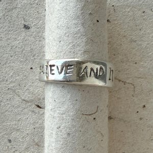 May include: A silver-colored band ring with a polished finish featuring the hand-stamped text "BELIEVE AND IT" clearly visible along the outer surface. The ring has a smooth, slightly irregular texture consistent with a handcrafted metal piece.